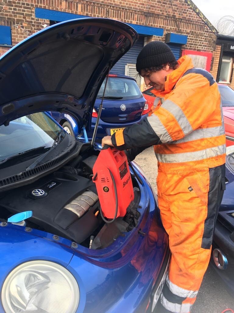 A Breakdown Man technician in hi-vis using a red jump pack on a blue VW Beetle during a roadside jump start.