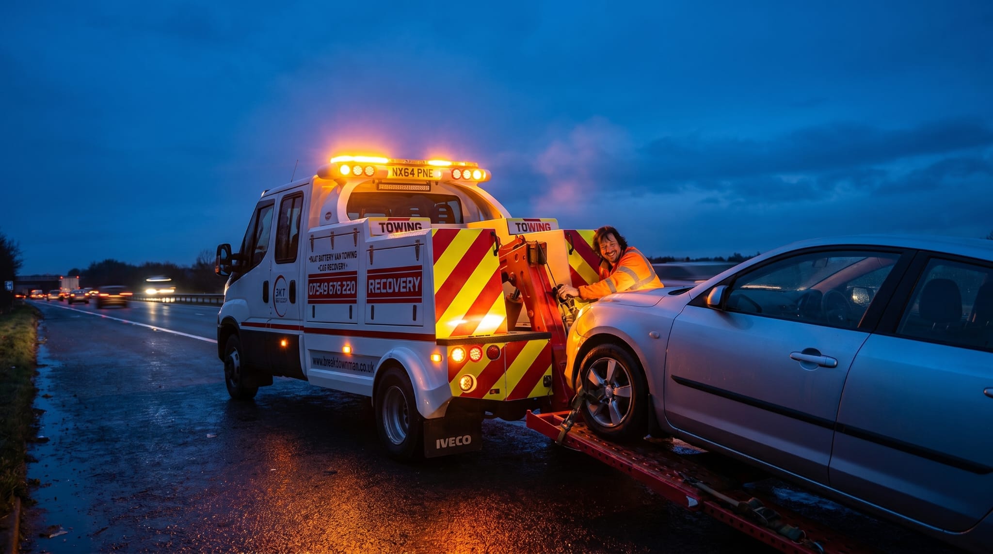 A Breakdown Man technician loading a silver car onto the Breakdown Man Iveco recovery truck at twilight on the motorway.