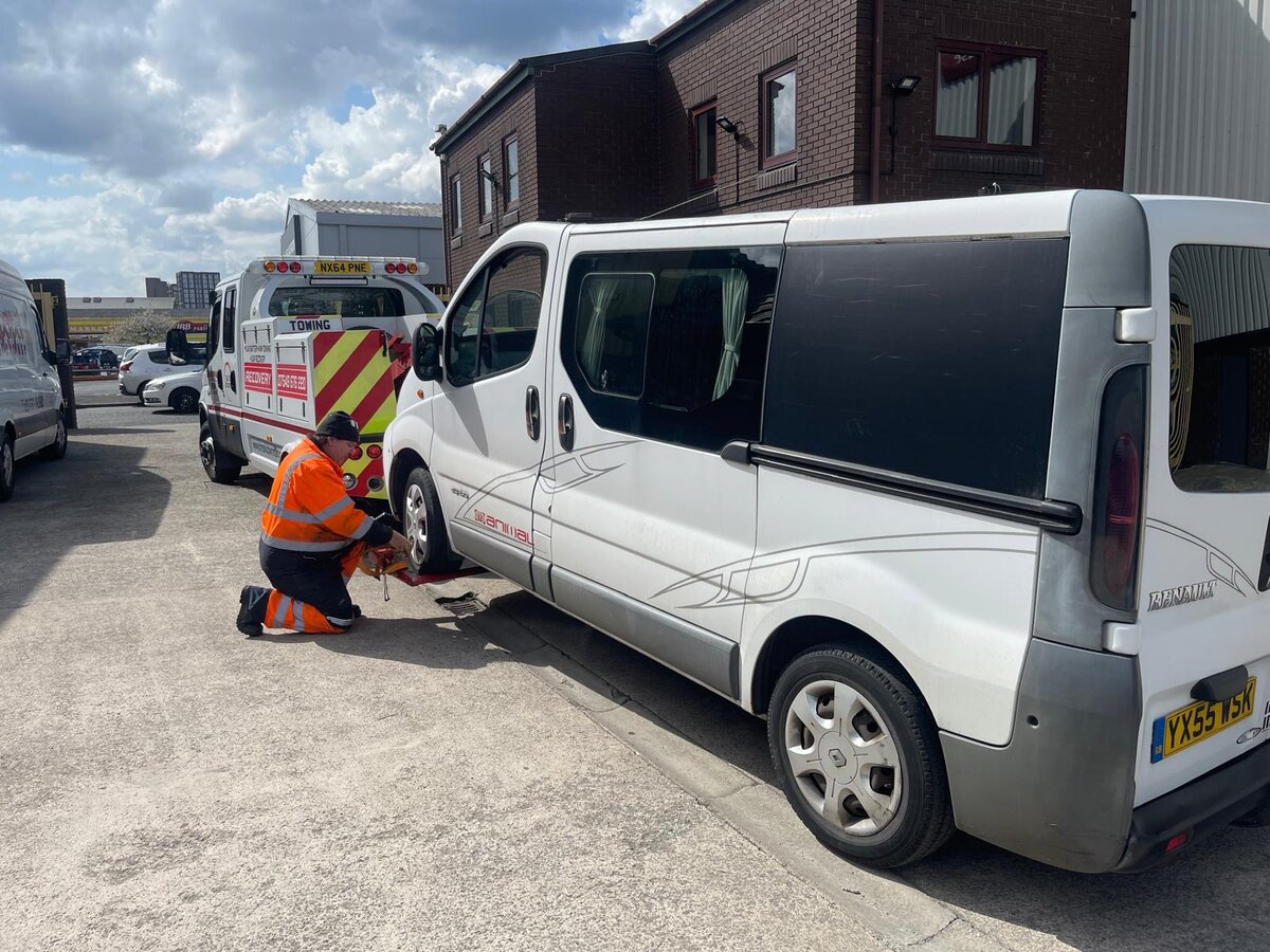 A Breakdown Man technician kneeling beside a silver Renault Trafic van, fitting recovery wheel-strap restraints at the depot before transport