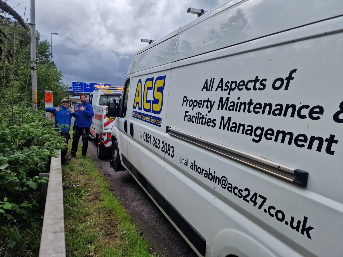 A Breakdown Man technician in hi-vis beside an ACS Property Maintenance white Citroen van being loaded onto a Breakdown Man recovery truck on a grass-verge roadside callout near the M57.