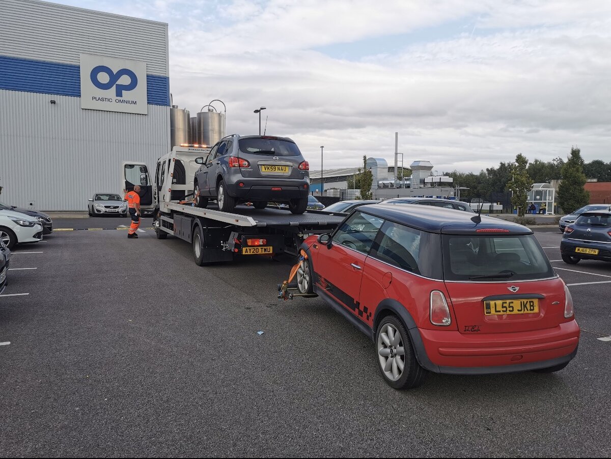 Breakdown Man recovering a grey Nissan Qashqai on the recovery truck with a red Mini towed behind, on an industrial site.